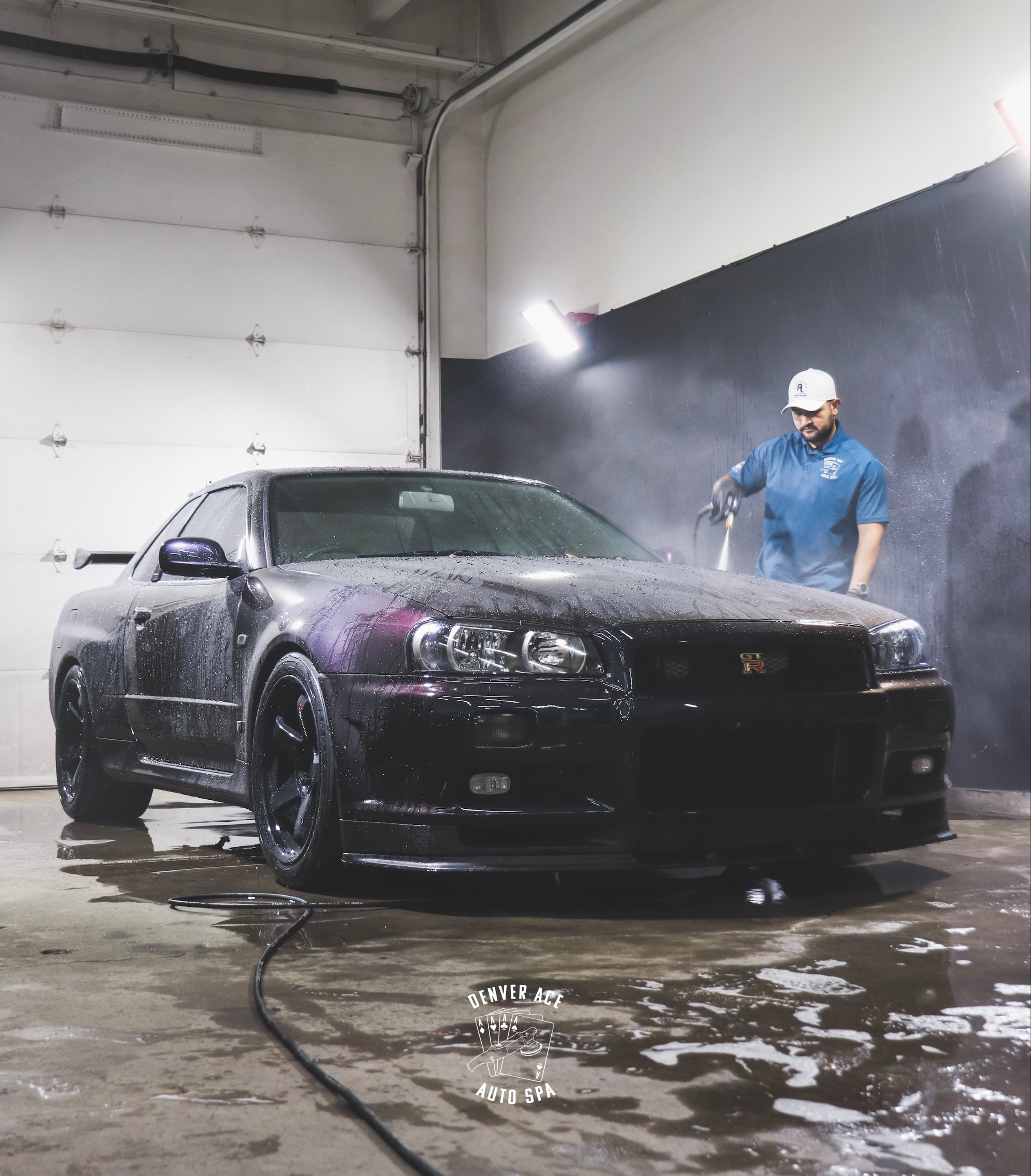 Technician pressure-washing a black Nissan Skyline GT-R in the Denver Ace Auto Spa shop bay, Denver Ace watermark visible