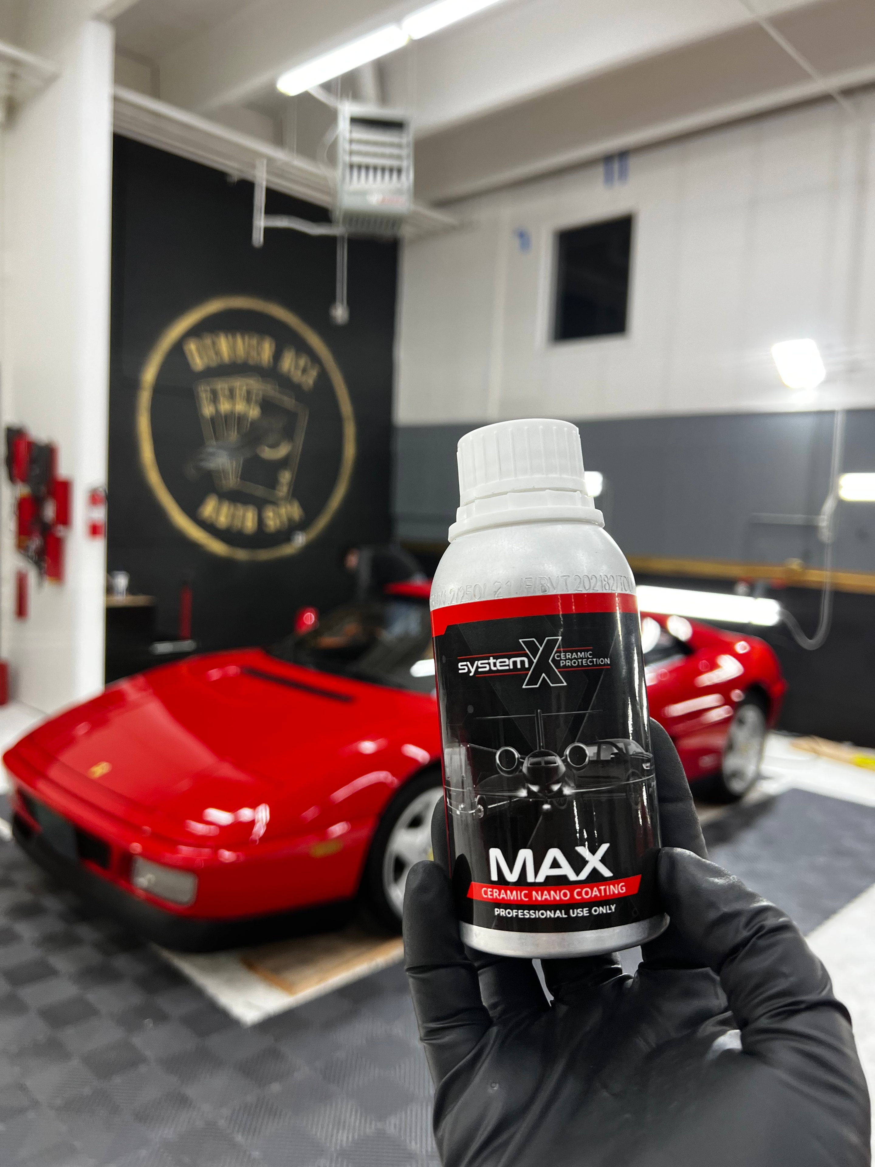 System X MAX ceramic nano coating bottle held up in the Denver Ace Auto Spa shop, red Ferrari and branded wall mural in background