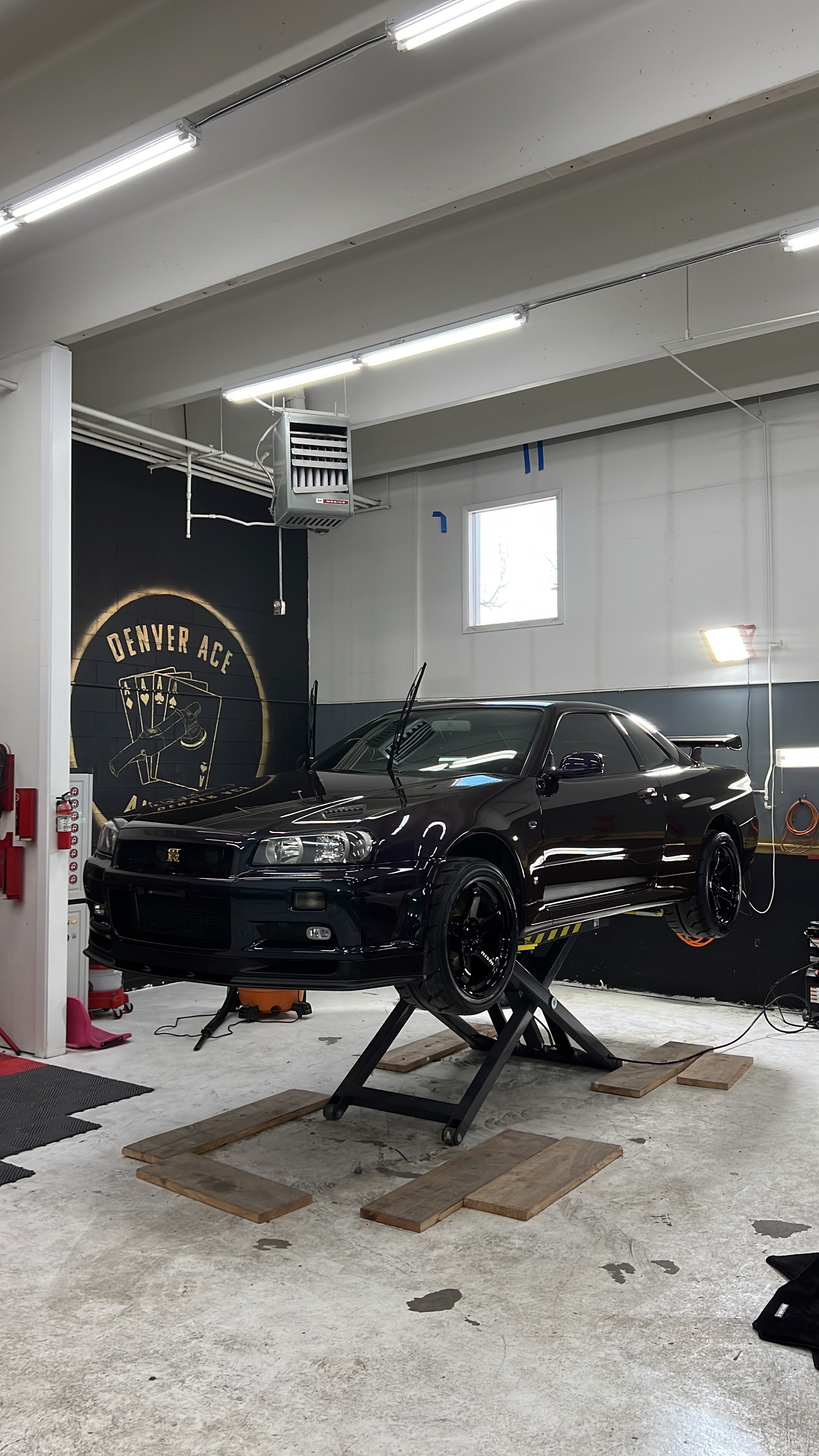 Black Nissan Skyline GT-R on a lift inside the Denver Ace Auto Spa shop, branded wall mural visible